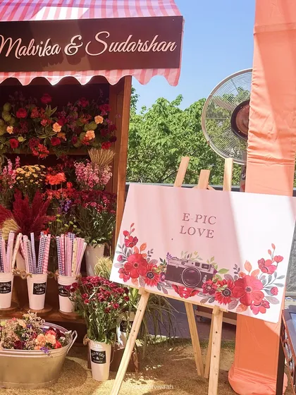 A personalized welcome sign with a vintage camera graphic, set beside the fresh flower stall, adding a cute and custom detail for the couple.