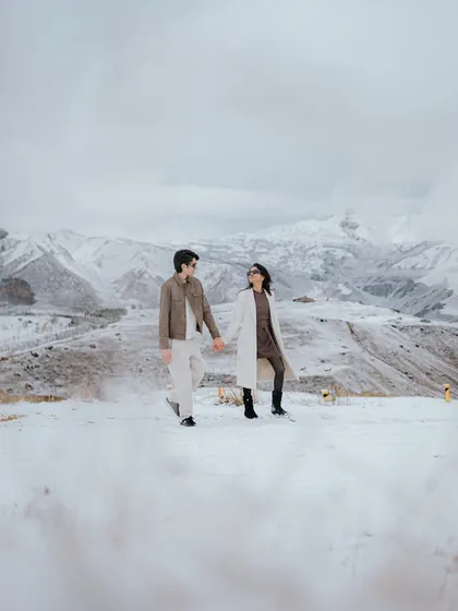 A wide shot capturing the vast, snow-covered mountains of Azerbaijan. The couple walks hand-in-hand, showcasing the grand scale and epic nature of a destination pre-wedding shoot.