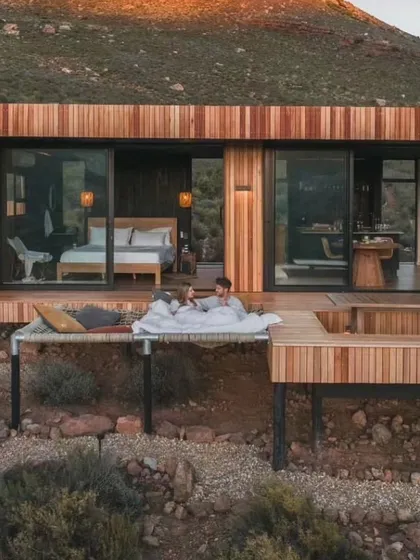 A couple enjoying the indoor-outdoor living experience of our modular cabin. The design features a bed that opens directly onto the deck, complete with a hammock and hot tub for a truly luxurious retreat.