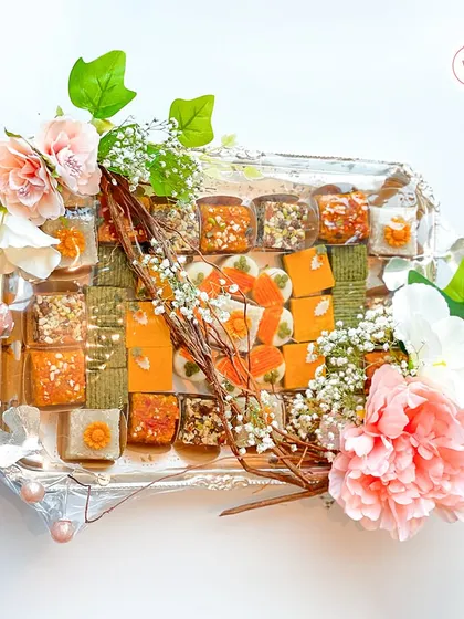 An exquisite mithai hamper on a silver tray, featuring a wide assortment of colorful Indian sweets and adorned with delicate pink and white flowers.