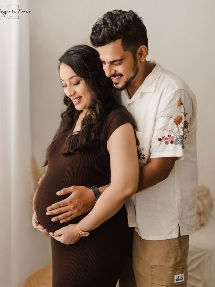 A gentle and loving maternity portrait. The husband wraps his arms around his wife, both looking down at the baby bump with sweet anticipation.