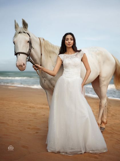 A full-length view of the model and horse on the beach. The natural, golden hour light creates a soft, romantic mood, perfect for a pre-wedding or conceptual fashion shoot.