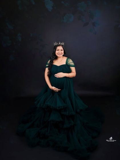 A regal solo portrait. The mom-to-be wears a crown and a gorgeous dark green gown, looking every bit the queen against a classic dark backdrop.