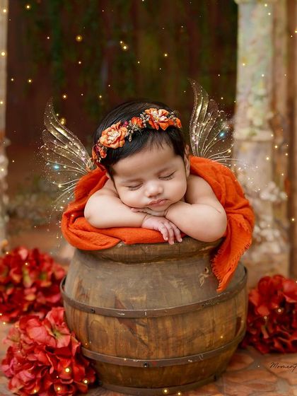 A little autumn fairy. With delicate wings and a floral crown, this newborn sleeps peacefully in a rustic barrel, surrounded by red flowers.
