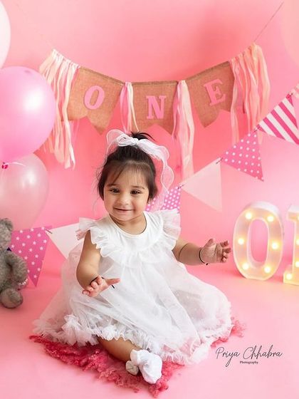 All set for her first birthday celebration! This little girl looks absolutely adorable in her white dress against the pink backdrop, ready for her party to begin.