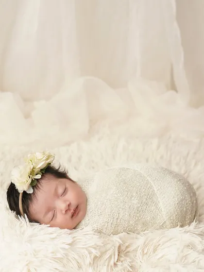 A full-body shot of the swaddled newborn, looking like a tiny, precious bundle on a cloud of white fur. The sheer curtain in the background adds a dreamy, ethereal quality.