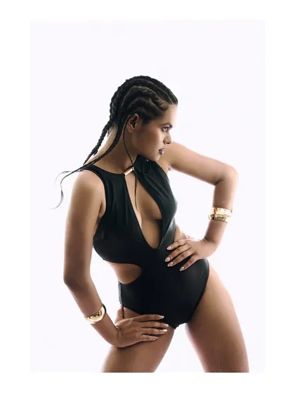 This dynamic pose adds energy to the minimalist setup. The model's posture highlights the cutouts of the swimsuit and the shine of the gold cuffs, demonstrating how to create visual interest even with a simple background.