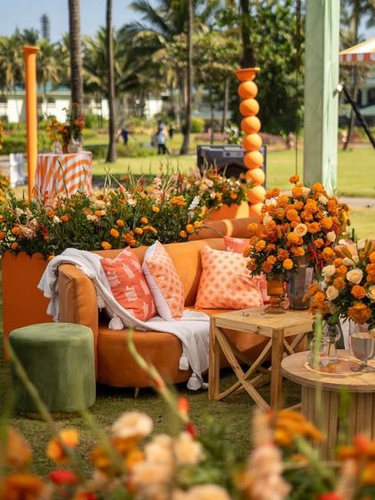 A comfortable and stylish lounge area at an outdoor haldi event. The orange sofa with patterned cushions is surrounded by marigold arrangements, creating a relaxed and festive corner.