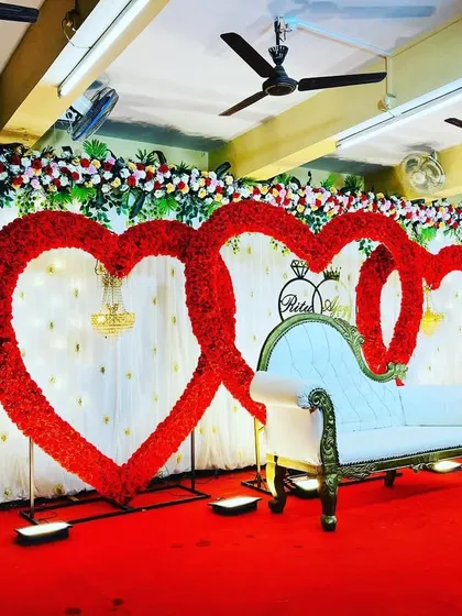 An elegant and romantic reception stage featuring two large, interlocking heart-shaped frames covered in red flowers. A classic white chaise lounge sits in front, set against a white curtain backdrop, perfect for a love-themed wedding.