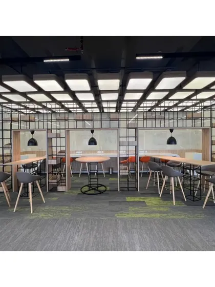 A semi-enclosed collaborative hub within the open office, featuring high tables and stools. The metal grid shelving provides a sense of separation without sacrificing the open feel, making it an ideal space for informal team meetings.