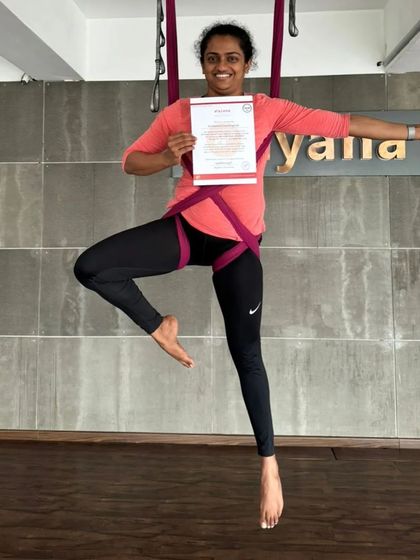 A student beams with joy, certificate in hand, as she masters a one-legged pose in the aerial hammock.