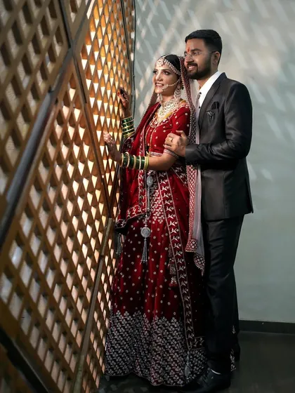 A stylish portrait of the couple after their ceremony. The bride in her red lehenga and the groom in his sharp suit stand by a decorative wall, blending tradition with modern elegance.