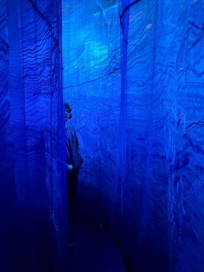 A visitor is enveloped by the deep blue, layered textiles of the 'Into The Deep' installation. This immersive experience is designed to evoke the feeling of being underwater, surrounded by the mysterious and haunting beauty of the ocean.