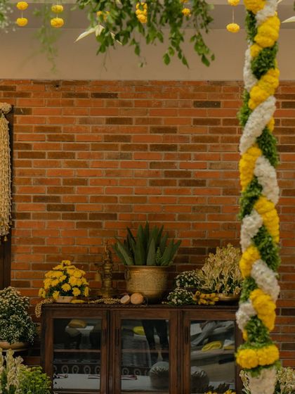 A detail shot of the mandap decor, showing a pillar wrapped in a spiral of marigold and jasmine garlands against a rustic brick wall. This contrast of textures adds a unique, modern touch to the traditional setup.