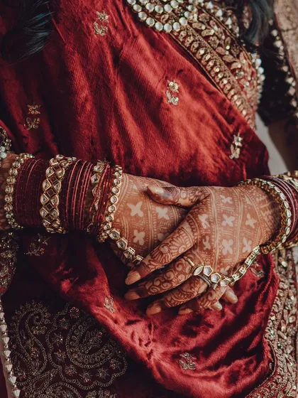 A detailed shot of a bride's hands, adorned with intricate henna and traditional bangles, resting on a rich velvet outfit. We believe the small details tell a big part of the wedding story.