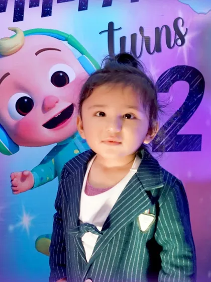 A close-up portrait of a toddler in front of his "turns 2" Cocomelon-themed birthday backdrop, capturing a sweet and direct gaze.