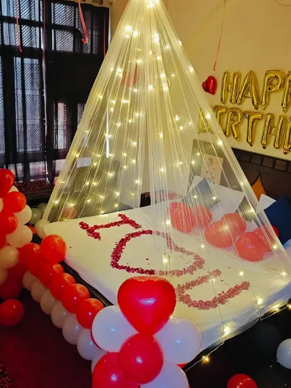 This birthday room setup combines a glowing canopy with a personal touch. Rose petals on the bed spell out "I ❤️ U," while "HAPPY BIRTHDAY" foil balloons hang above, all in a classic red and white theme.