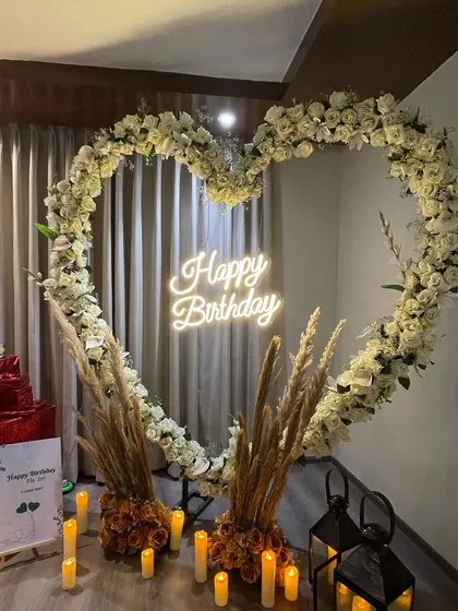 A romantic birthday decoration centered around a large heart-shaped arch made of white flowers. A 'Happy Birthday' neon sign and warm candlelight create a soft and elegant ambiance for the celebration.