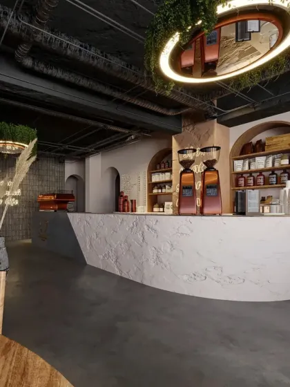 The cafe's counter is designed with a rough, textured finish, contrasting with the sleek coffee machines. The hanging greenery and warm lighting add to the organic and welcoming feel.