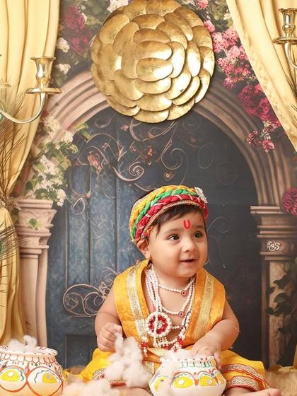 Our little Kanha sits amidst a grand, palace-like setting with golden curtains and candelabras. The entire setup is designed to create a regal and divine portrait.