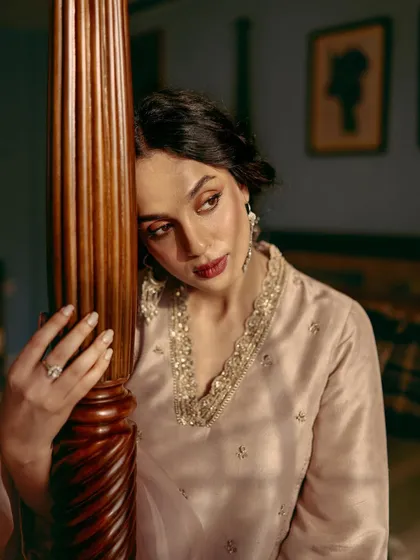 A pensive portrait for AR by Rhea Kapoor. The model leans against a wooden bedpost, and the warm, soft lighting creates an intimate and cinematic mood.