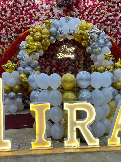 A stunning first birthday stage with a red flower wall, a gold and silver balloon arch, and large marquee letters spelling 'IRA'.