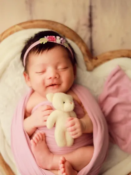 That fleeting, precious newborn smile. We live for these moments of pure joy. This little one is settled comfortably in our heart-shaped wooden prop, wrapped in a soft pink swaddle for a classic and heartwarming portrait.