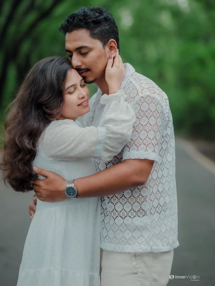 A close, tender embrace on a quiet road. The groom kisses his partner's forehead, a classic gesture of love and affection captured beautifully in the soft, natural light.
