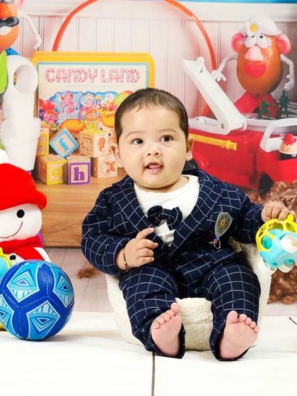 A baby boy in a dapper plaid suit sits among a sea of toys, looking calm and content.