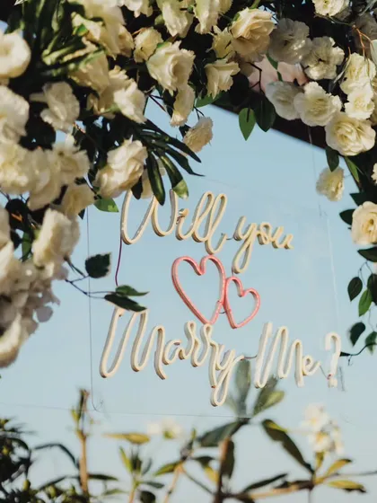 A close-up detail of the custom "Will you Marry Me?" sign from our rustic white floral arch setup. The clear acrylic and delicate script add a modern touch to the romantic floral arrangement.