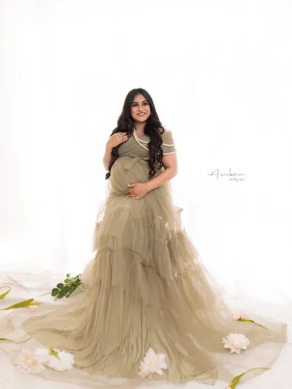 A full-length shot showing the beautiful tiered design of the olive gown. The simple, bright background keeps the focus on her radiant smile and the elegant dress.
