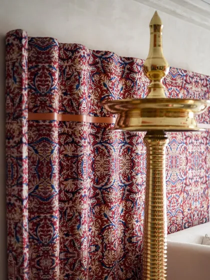 A magnificent 6-foot tall brass diya is strategically placed against the custom Kalamkari art piece, casting a warm glow and highlighting the beauty of South Indian heritage.