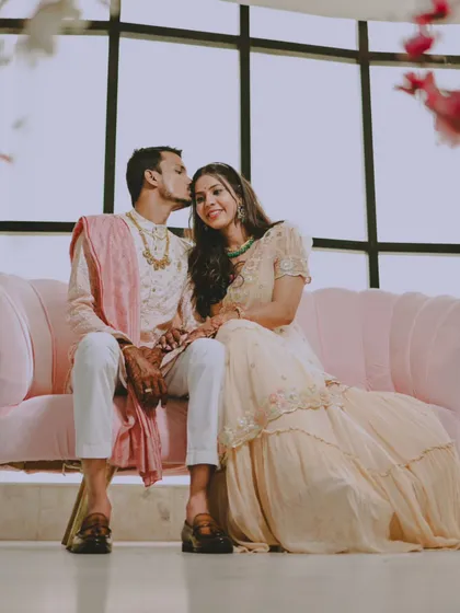 A full-length portrait of the couple on a pink sofa, with the groom giving his fiancée a gentle kiss on the forehead.
