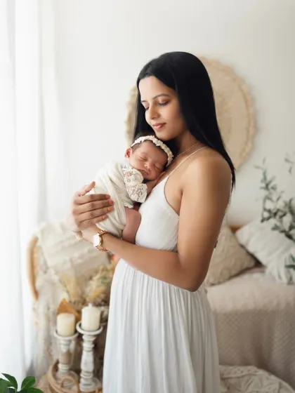 A mother in a beautiful white dress holds her sleeping newborn daughter. The soft, natural light highlights the peacefulness of this moment.