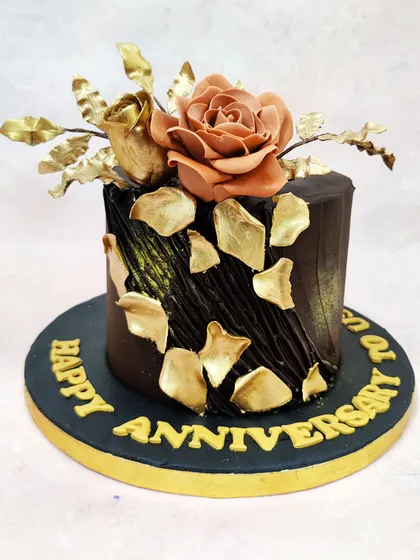 This anniversary cake is a statement of opulent romance, featuring a rich, dark chocolate exterior with a textured finish. A handcrafted sugar rose in a muted blush tone is complemented by gilded leaves and shimmering gold leaf fragments, creating a luxurious and timeless design for celebrating a special milestone.