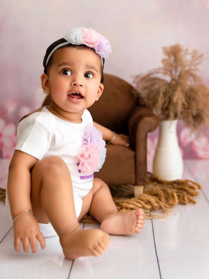 A baby girl sits on the floor next to a miniature armchair in a pastel-themed setup.