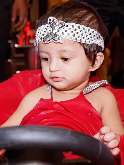 A close-up of a baby girl in a red dress sitting in a toy car, a popular prop for first birthday photoshoots.