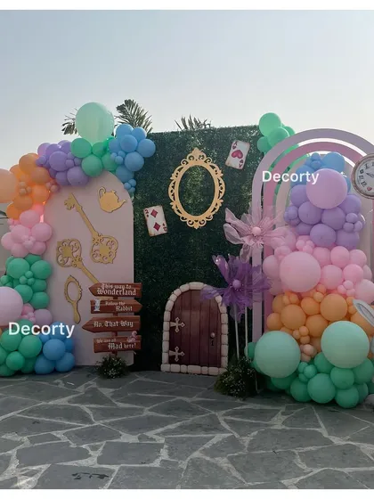 An "Alice in Wonderland" backdrop featuring a grass wall, a small door, and colorful balloon arrangements.