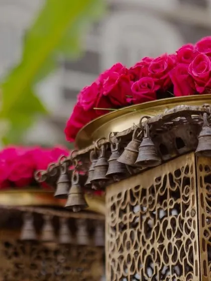 A close-up of a custom-made brass lantern with intricate 'jaali' work and hanging bells, topped with a vibrant arrangement of pink roses.