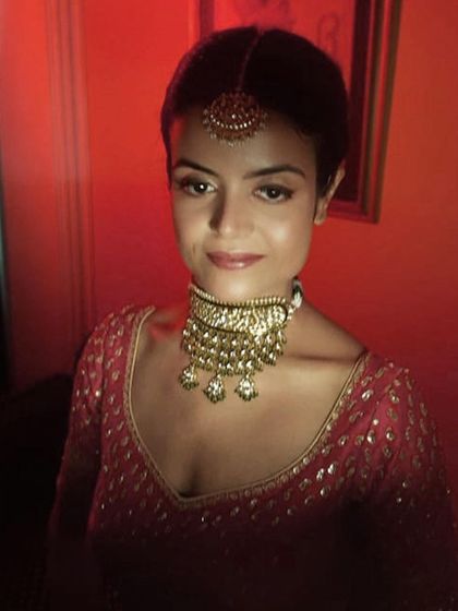 A close-up portrait of a bride under dramatic lighting. The focus is on her glowing skin and the subtle shimmer of her makeup, paired with a traditional gold choker and maang tikka.