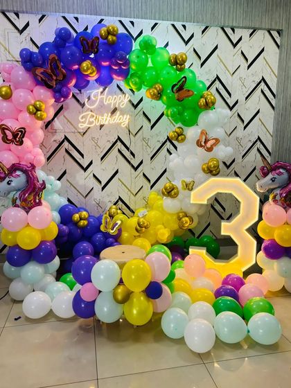 A whimsical unicorn and rainbow theme for a third birthday. The colorful balloon arch is decorated with unicorn and butterfly foils, paired with a neon sign and a light-up number '3'.