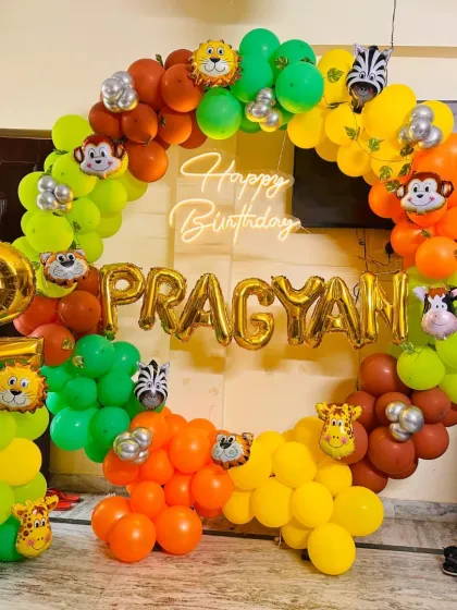 A closer look at a jungle safari theme, perfect for a second birthday. This decoration features a full circular balloon arch in vibrant greens, yellows, and oranges, dotted with cute animal foil balloons. The design is personalized with the child's name, 'PRAGYAN', in gold foil letters and a central 'Happy Birthday' neon sign.