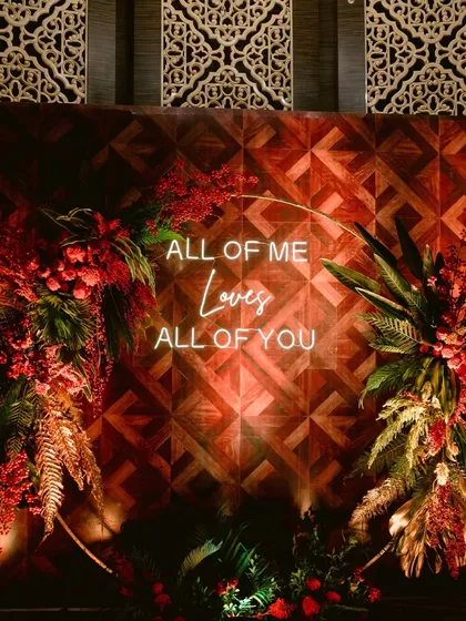 A romantic neon sign reading "All of me Loves all of you" serves as the perfect photo backdrop for a Sangeet night. The sign is set against a rich, wooden-paneled wall and framed with red and gold foliage.
