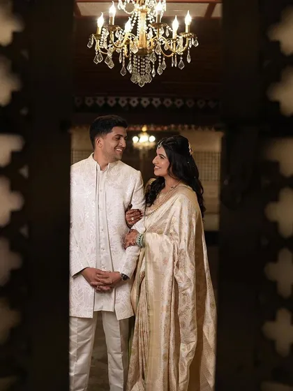 A regal portrait of a couple. Her elegant off-white saree and soft, wavy hair create a look of timeless sophistication, perfectly captured under a grand chandelier.