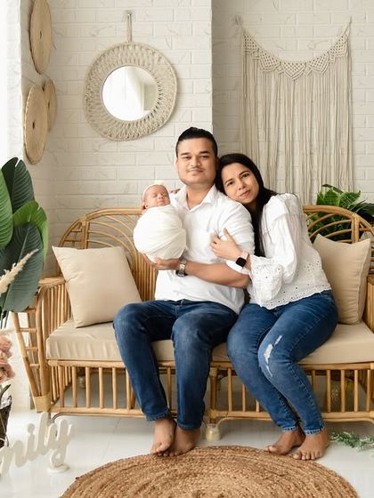 A full family portrait in our popular boho-themed studio. The family is casually posed on the rattan sofa, creating a relaxed and natural lifestyle photograph with their newborn.