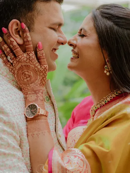 A tender close-up of a woman gently touching her partner's face, their noses almost touching. The shot beautifully details her intricate henna design, capturing the intimate details and emotions of a pre-wedding celebration.
