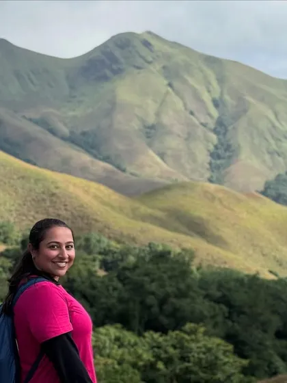 A smiling trekker poses with the stunning Kudremukha mountain range behind her. The views are simply spectacular.