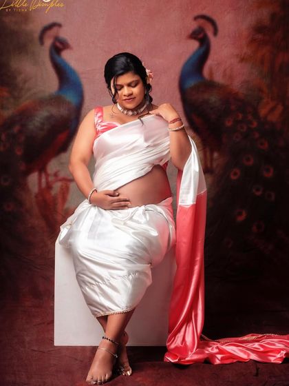 A beautiful seated portrait of a mom-to-be in a white satin saree. The peacock-themed backdrop adds a touch of traditional elegance.