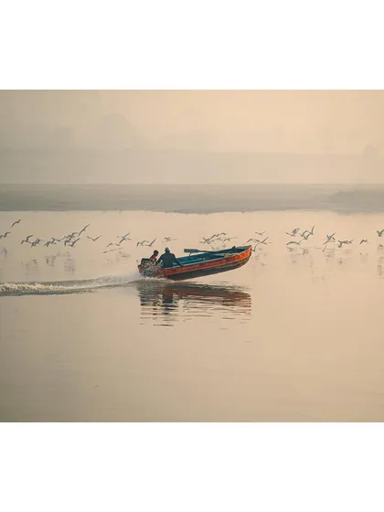 A speedboat cuts across the calm surface of the Yamuna Ghat, with birds taking flight in its wake. The misty background and soft light give the scene an ethereal quality.