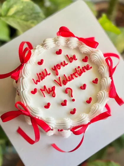 The sweetest way to ask. This Valentine's cake features a clean white top with red script and hearts, framed by a piped border and bright red ribbons.